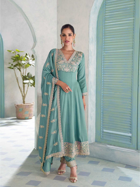 A woman models a mint green traditional Indian salwar kameez suit with intricate gold embroidery, standing in a courtyard with arches and shutters.