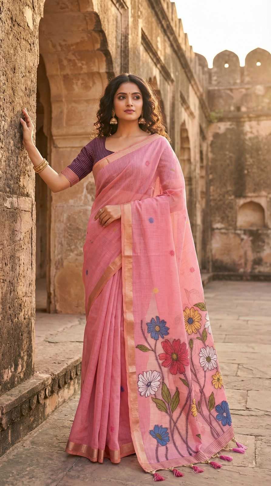 Woman in a pink saree with floral patterns standing in front of an ancient architectural structure.
