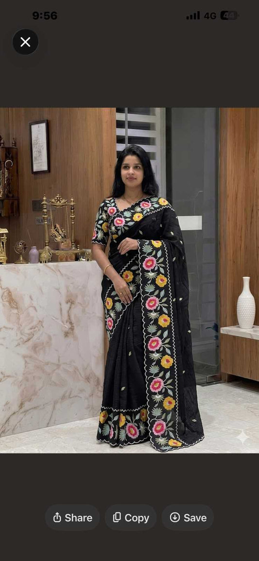 A woman wearing a black saree with colorful floral embroidery, standing in an indoor setting with a marble counter and wooden wall in the background.