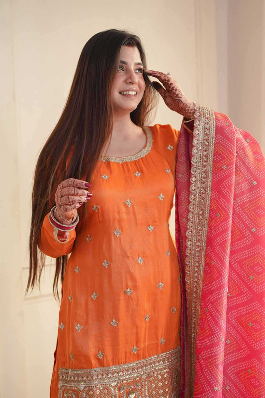 A woman wearing a bright orange embroidered kurta with a pink patterned dupatta draped over her shoulder, standing against a plain beige wall.
