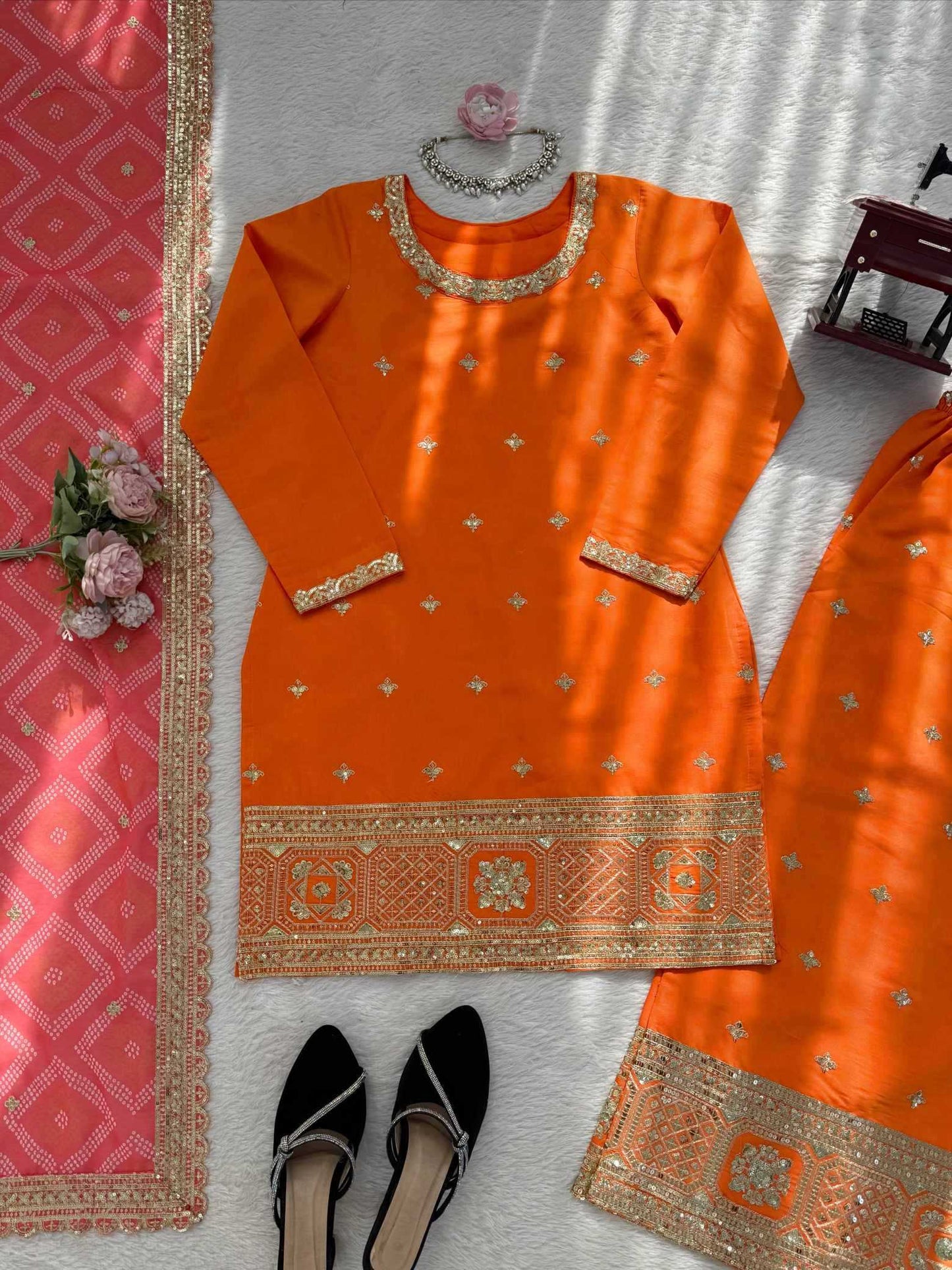 An orange traditional kurta set with gold embroidery, laid flat on a white textured surface with a pink dupatta, black flats, and decorative flowers.