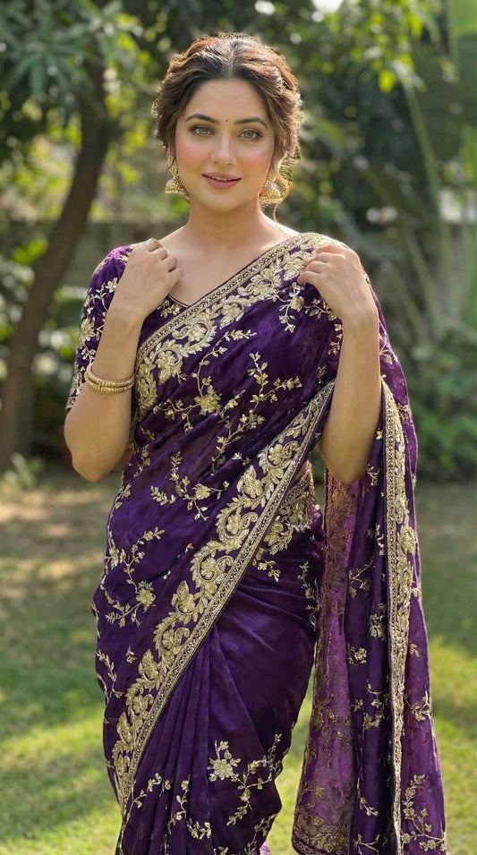 A woman wearing a purple sari with intricate gold embroidery, standing outdoors in a park-like setting with trees in the background.