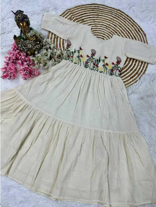An off-the-shoulder cream-colored cotton dress with colorful floral embroidery across the chest, styled on a white faux fur surface with a woven straw hat, pink flowers, and a small Buddha statue.