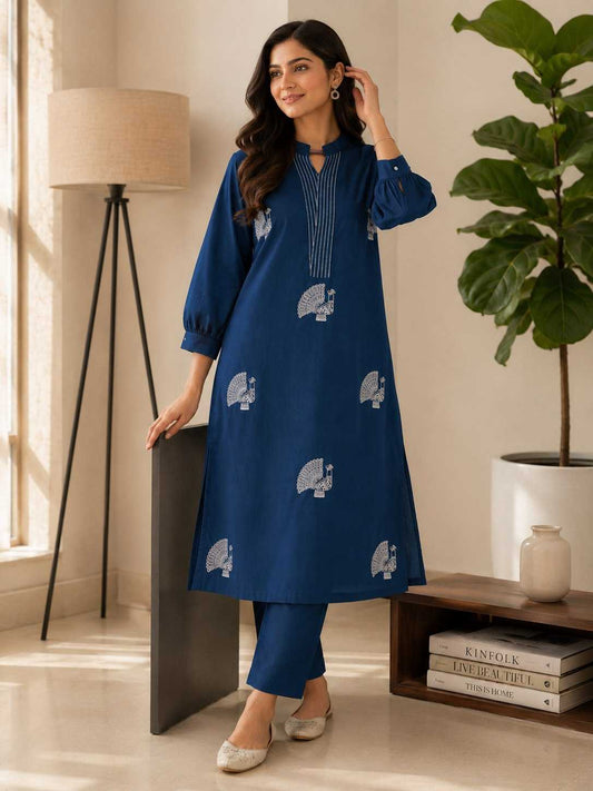 Woman in a blue dress with white patterns standing in a room with a lamp and plant.