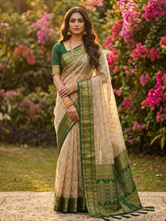 Woman in a green and beige saree standing in a garden with pink flowers.