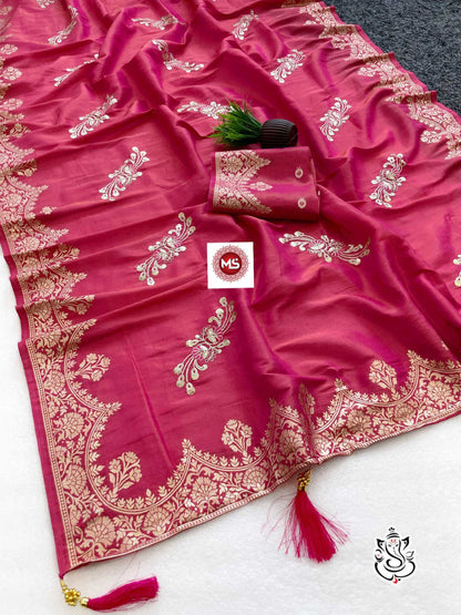 A magenta pink silk saree with intricate gold zari floral motifs and a wide decorative border, laid out on a white surface with a matching folded fabric piece and tassels.