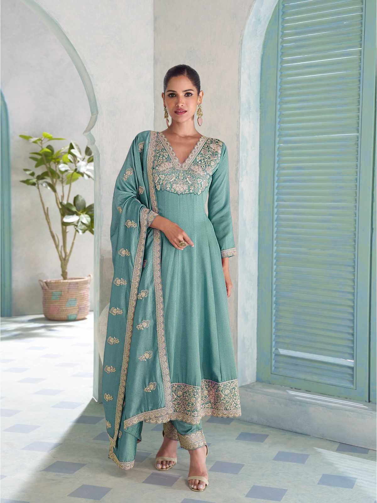 A woman models a mint green traditional Indian salwar kameez suit with intricate gold embroidery, standing in a courtyard with arches and shutters.