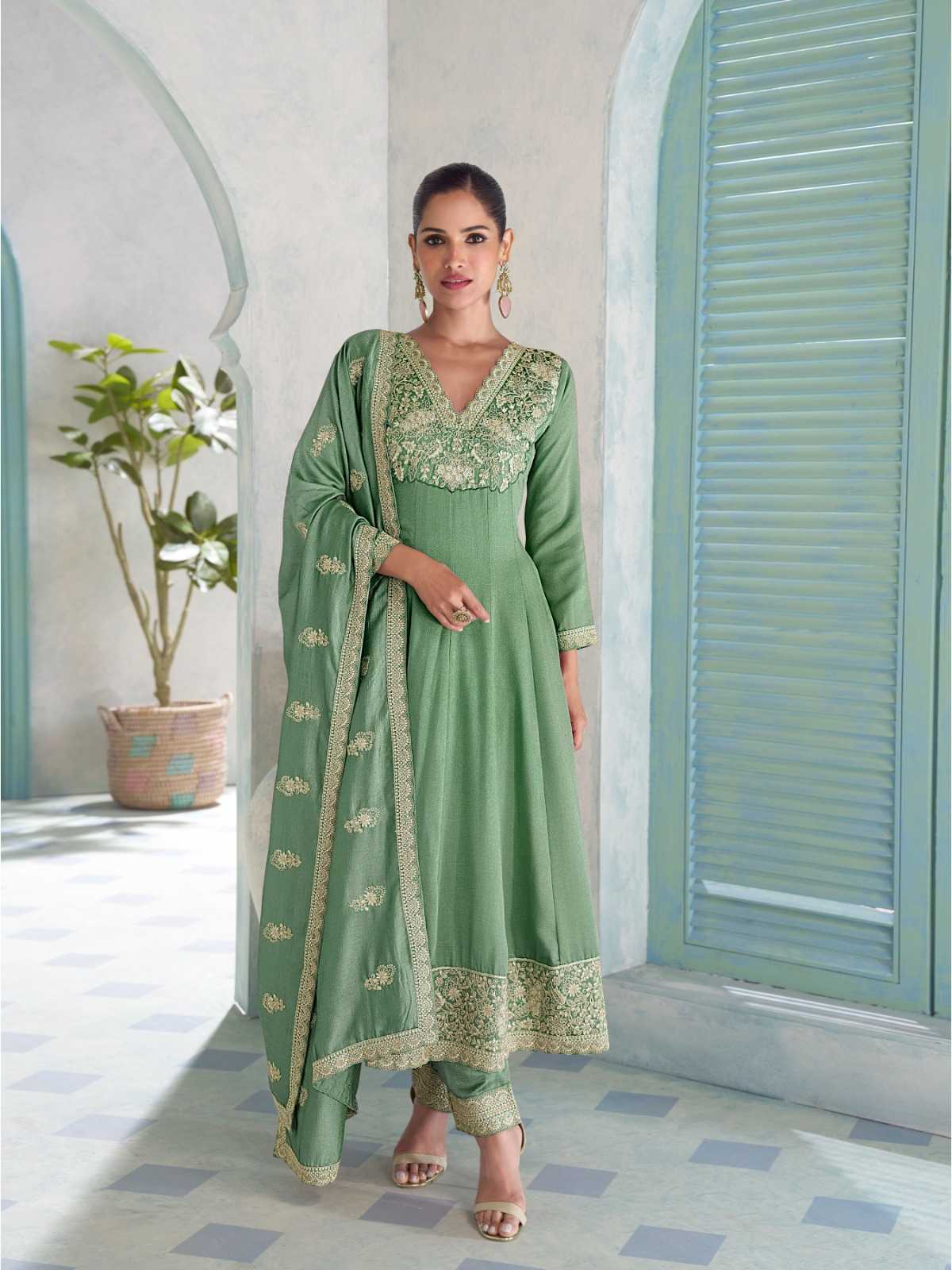 Woman in a green traditional outfit standing in a room with light blue shutters and a plant.