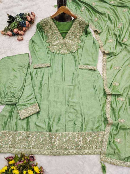 A sage green three-piece salwar kameez suit set with intricate gold and silver embroidery, laid out on a white textured background with decorative flowers.