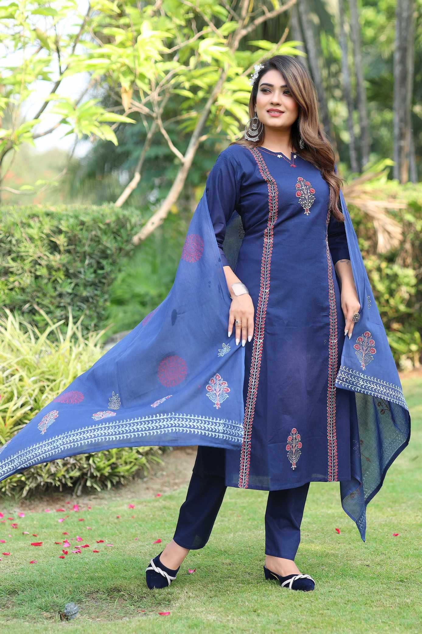 Woman in a blue traditional outfit with floral patterns standing outdoors on grass.