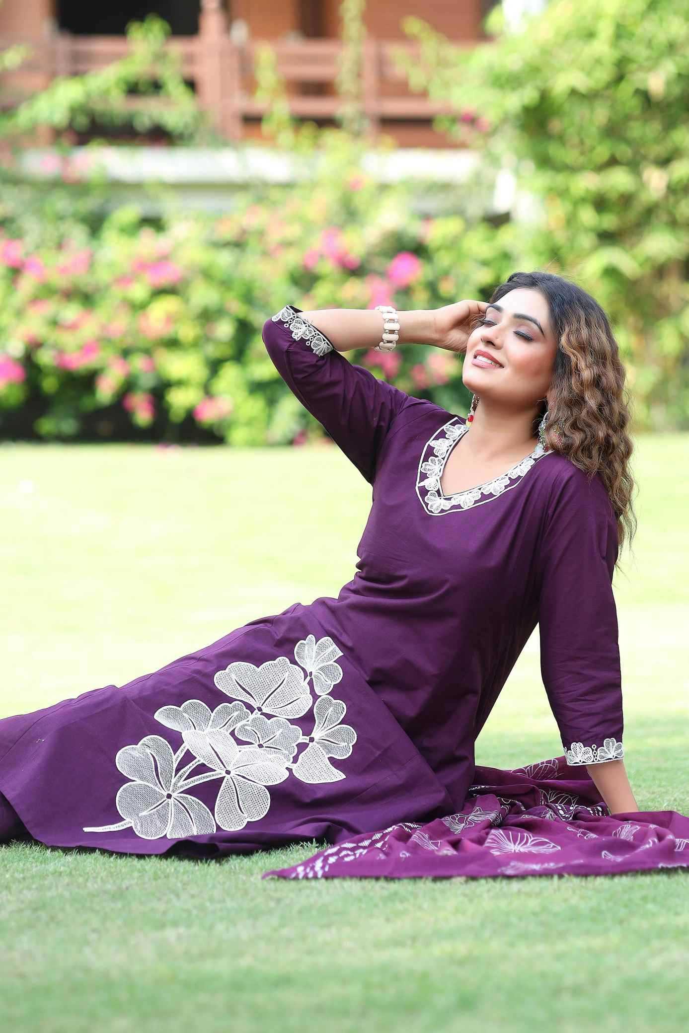 Woman in a purple dress with white floral patterns sitting on grass with flowers and a building in the background