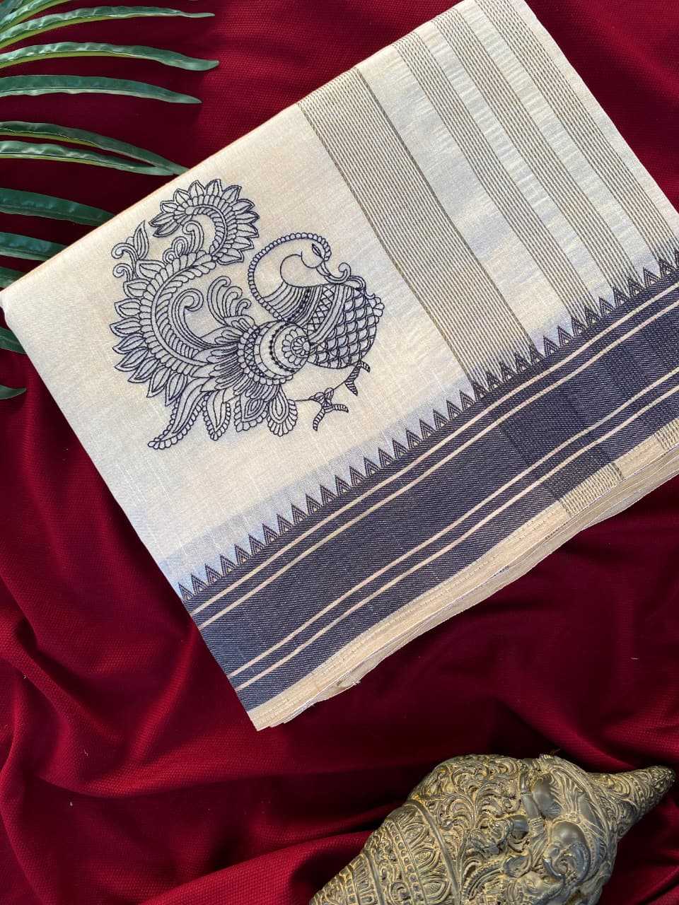 A folded cream-colored cotton saree or fabric piece with a navy blue peacock block print and striped borders, displayed on a deep red velvet fabric background with decorative green leaves and a metallic object.
