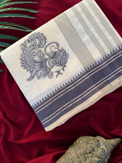 A folded cream-colored cotton saree or fabric piece with a navy blue peacock block print and striped borders, displayed on a deep red velvet fabric background with decorative green leaves and a metallic object.