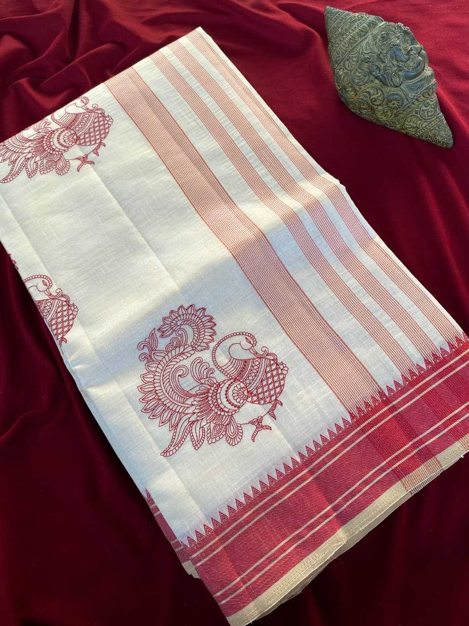 A white cotton saree or fabric piece with pink stripes, red paisley motifs, and a red-and-white patterned border, laid flat on a deep red velvet surface with a small green decorative object in the corner.