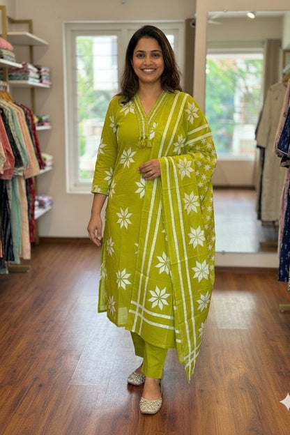 A woman modeling a lime green three-piece salwar kameez set with white floral prints, standing inside a clothing boutique.