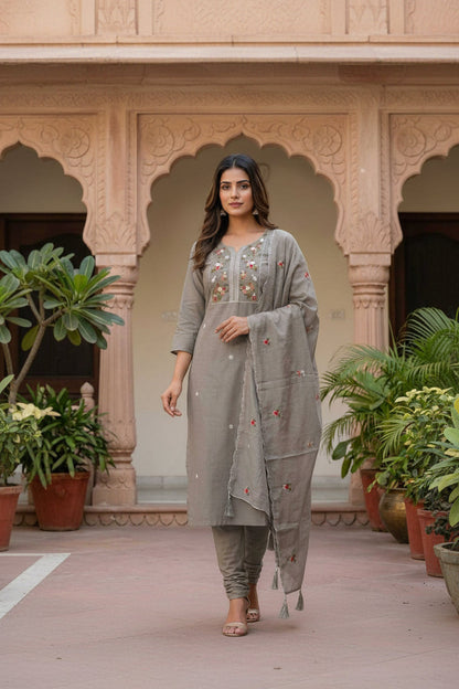 Woman wearing a gray embroidered outfit standing in front of an architectural background with plants.