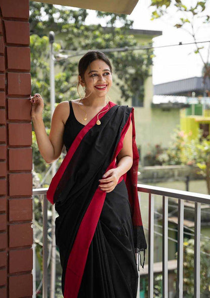 Woman in a black saree with red border standing on a balcony.