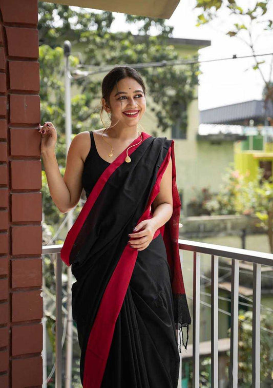 Woman in a black saree with red border standing on a balcony.
