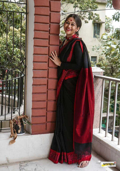 Woman in a black and red saree standing against a brick wall with greenery in the background