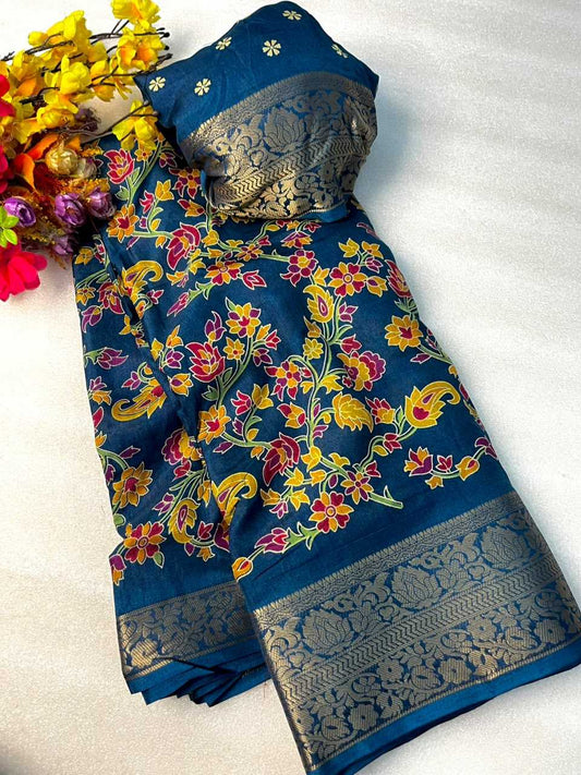 A folded blue saree with a vibrant floral and paisley print in yellow, red, and green, featuring a wide metallic gold border, displayed on a white fabric background next to yellow flowers.
