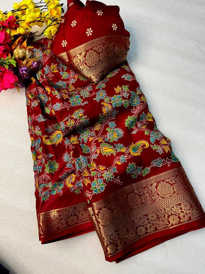 A folded deep red silk saree with intricate floral and paisley motifs in blue, green, and yellow, featuring a wide gold zari border, displayed on a white surface next to decorative flowers.