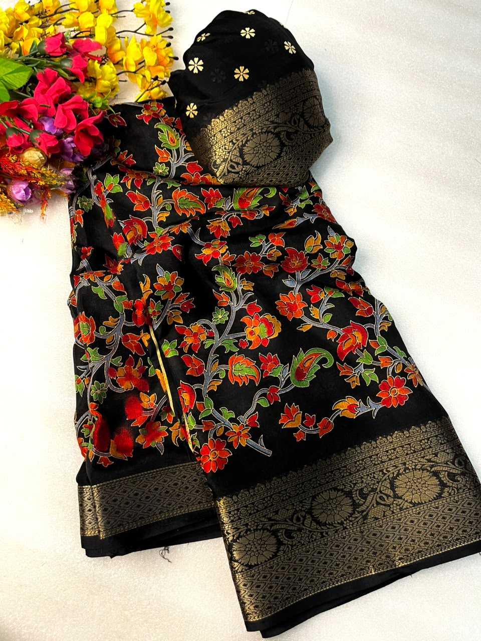 Black cotton saree fabric with a vibrant red, orange, and green floral pattern, featuring wide gold zari borders and a matching black dupatta with gold floral details, displayed on a white background next to artificial flowers.