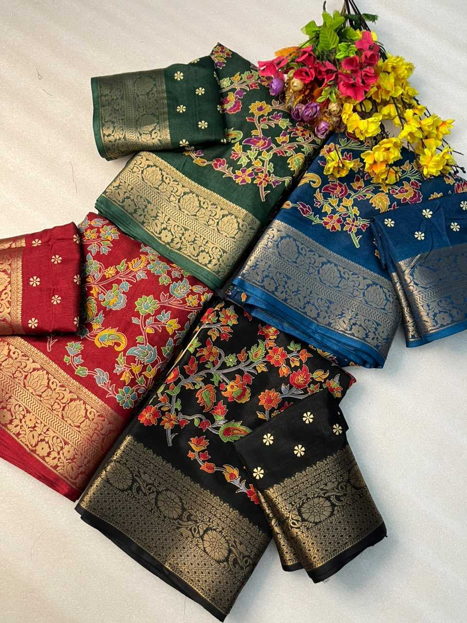 A flat-lay display of six traditional Indian silk sarees with intricate floral patterns and metallic zari borders, arranged on a light fabric background with a decorative flower bouquet.