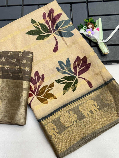 A beige cotton saree with large multicolor leaf motifs in green, purple, and gold, featuring a wide metallic gold border with elephant and peacock designs. A matching folded portion of the saree is visible on the left, and a small decorative flower arrangement sits on a dark slatted surface in the upper right.