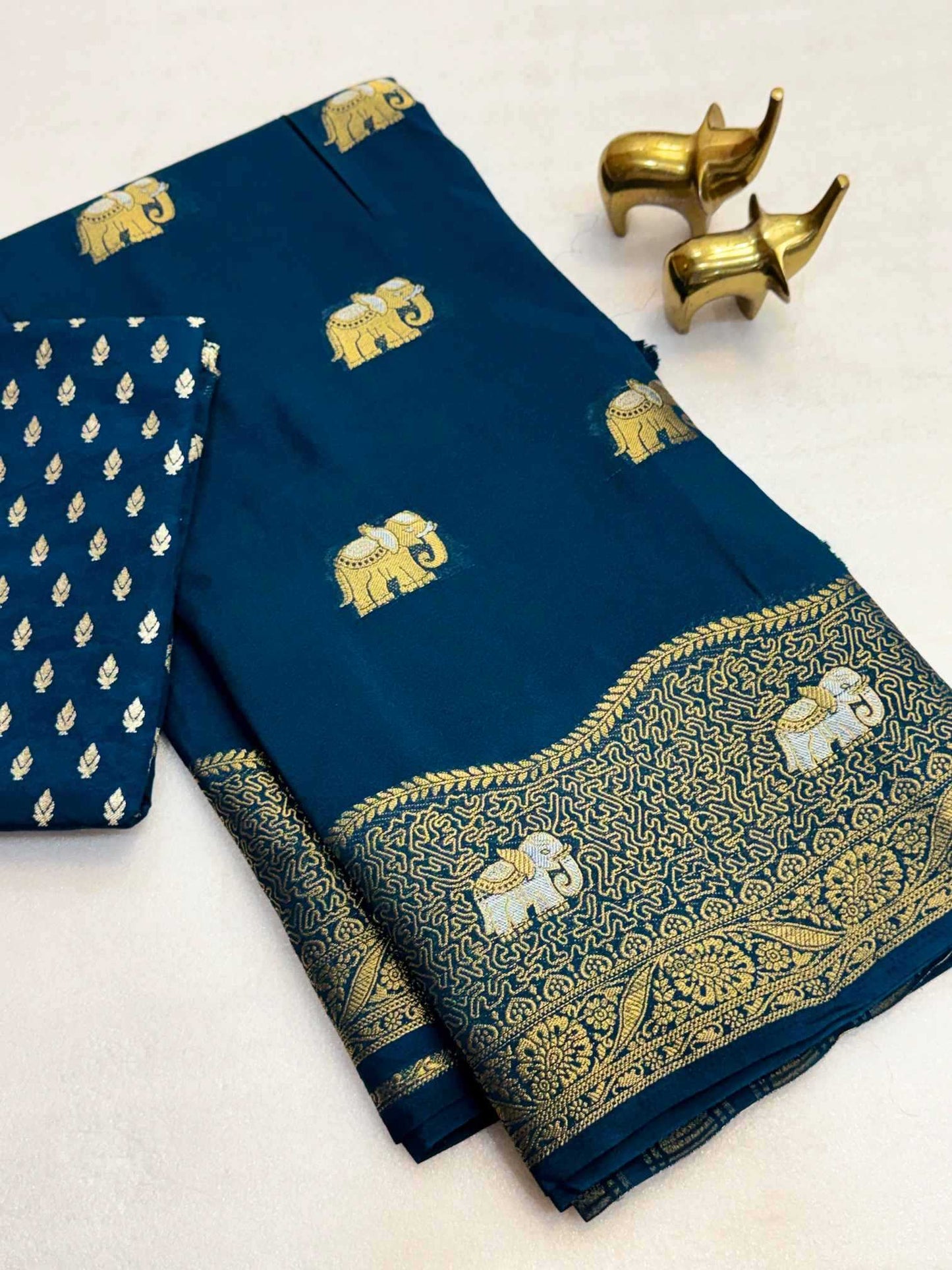 A folded navy blue fabric garment, likely a sari or dress material, featuring gold elephant motifs and an ornate gold border, with a matching patterned fabric piece folded over on the left. Two small gold elephant figurines are placed on a light background.