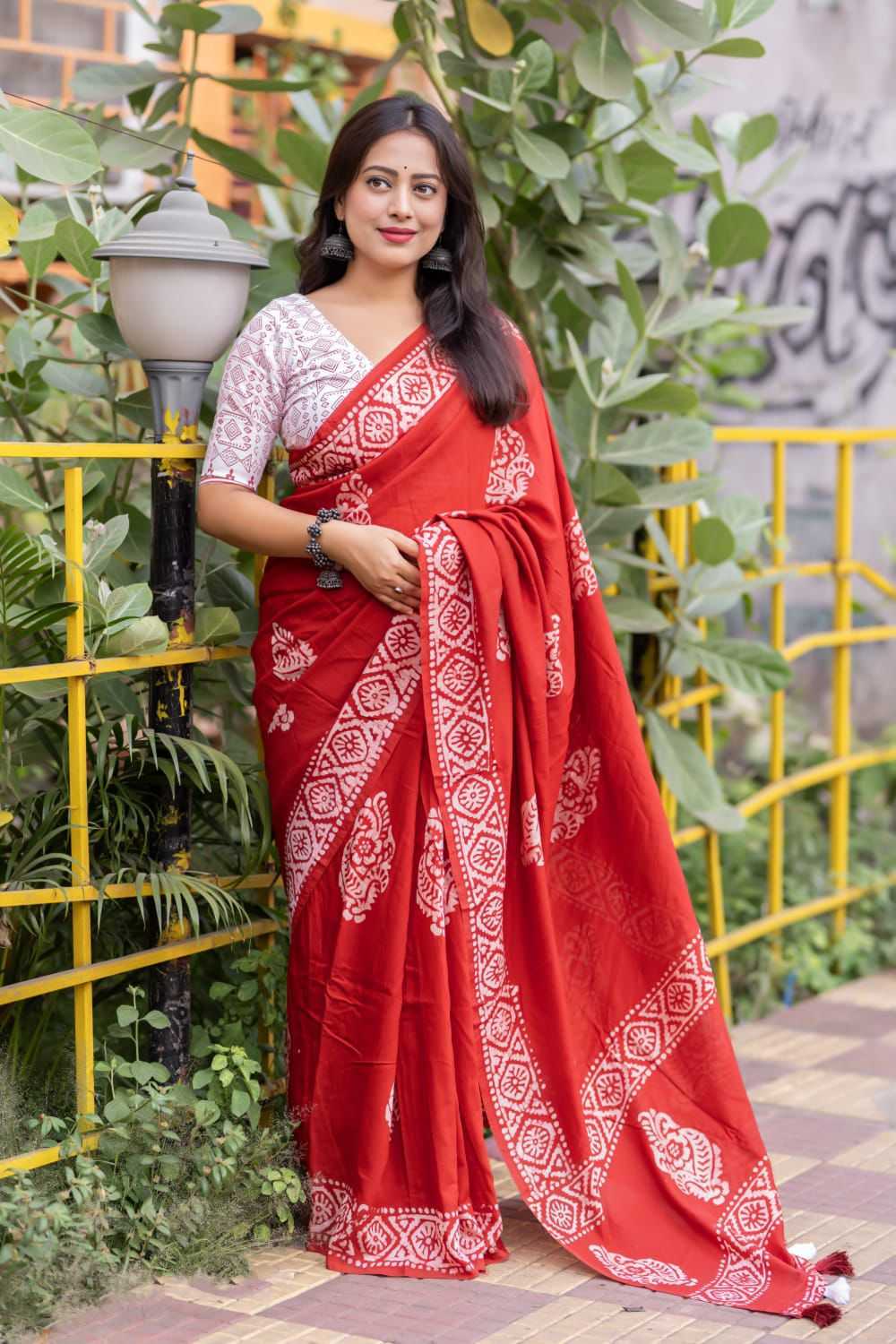 Woman wearing a red saree with white patterns standing outdoors near greenery.