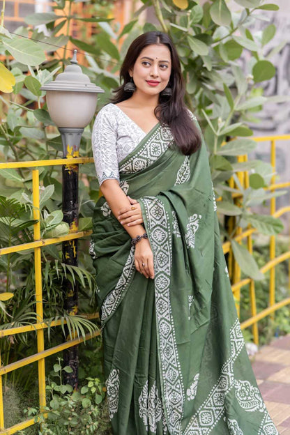 Woman wearing a green saree with white patterns standing outdoors with plants and a yellow railing in the background.