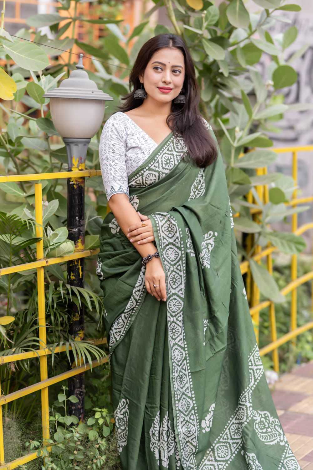 Woman wearing a green saree with white patterns standing outdoors with plants and a yellow railing in the background.