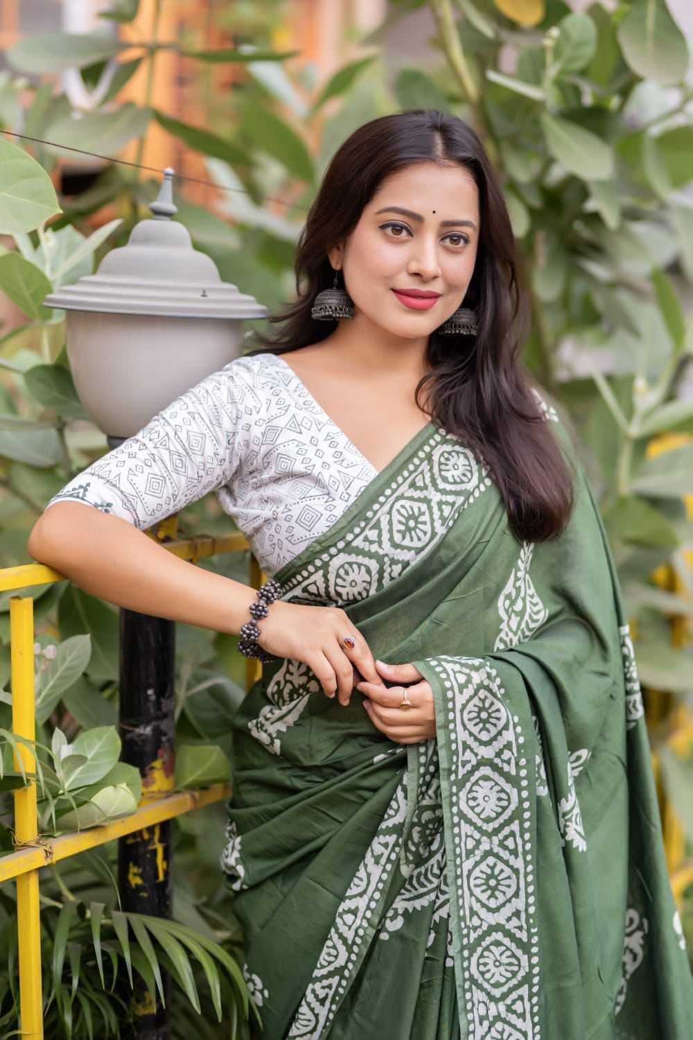 Woman in a green saree with white patterns standing outdoors among plants.