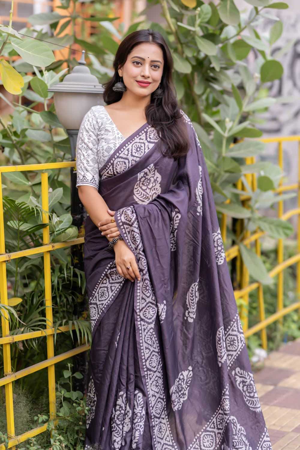 Woman wearing a purple saree with white patterns standing outdoors with greenery in the background