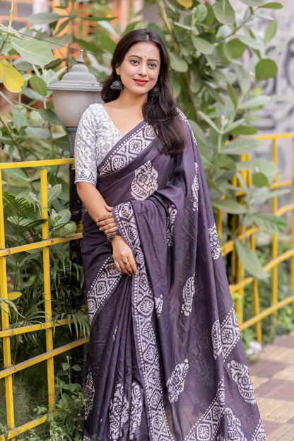 Woman wearing a purple saree with white patterns standing outdoors with greenery in the background