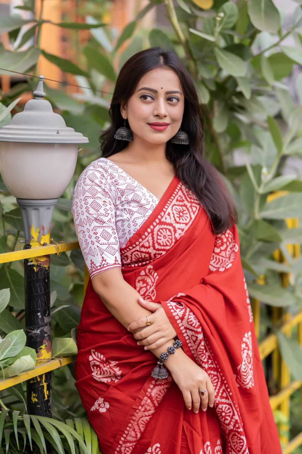 Woman in a red and white saree standing outdoors with greenery in the background