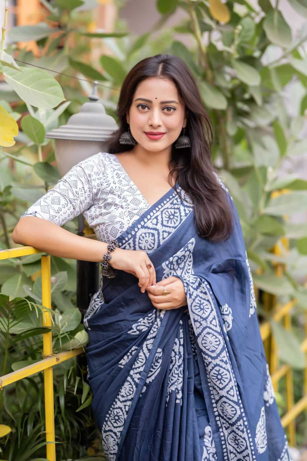 Woman wearing a blue saree with white patterns against a green foliage background