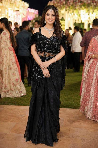 Woman in a black saree with floral patterns at an event with decorative lights and people in the background.