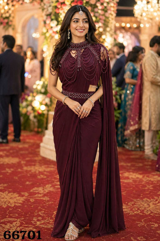 Woman in a maroon saree with intricate designs at an event with floral decorations and people in the background.