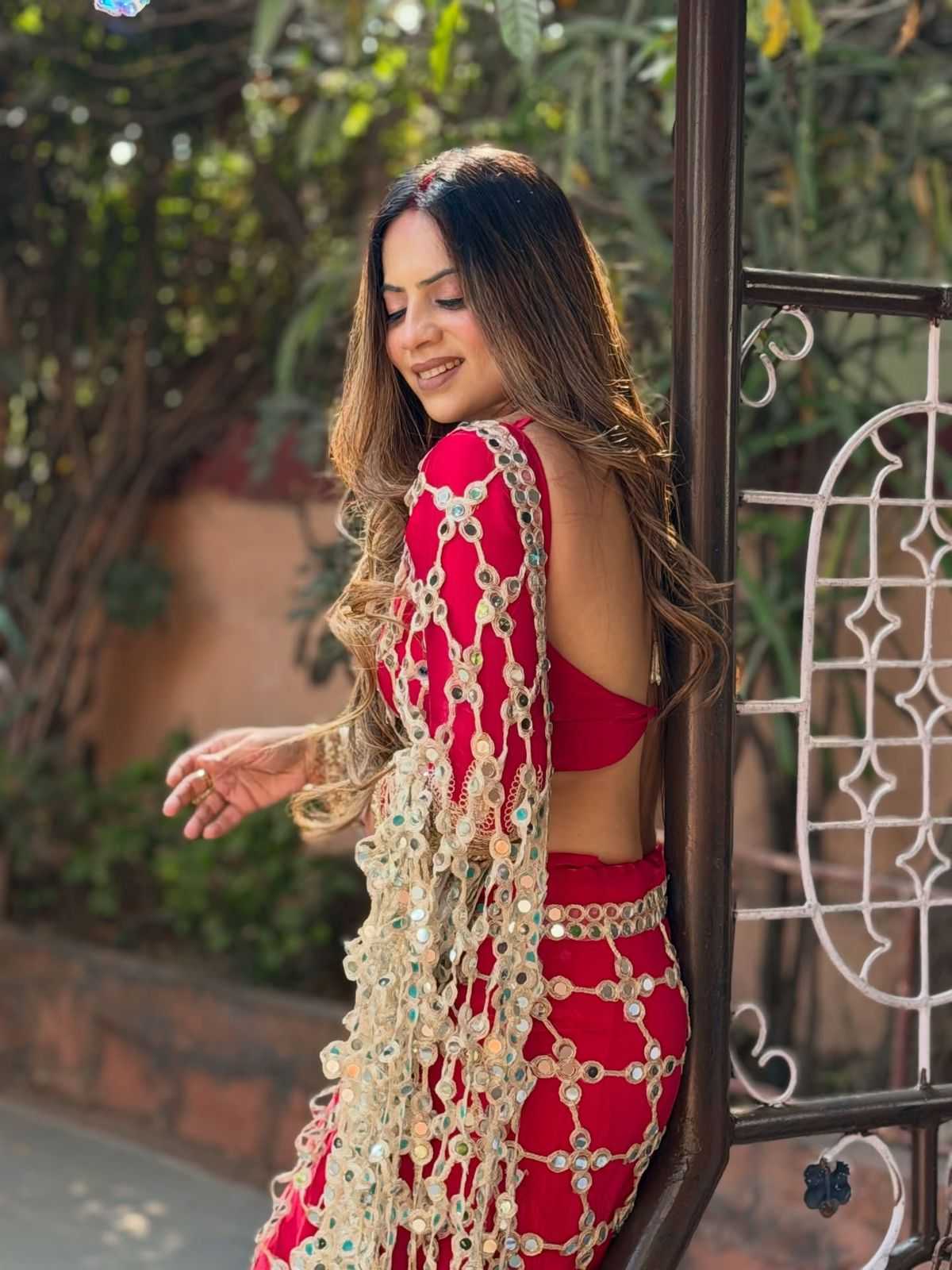 Woman in a red and gold traditional outfit standing outdoors with greenery in the background