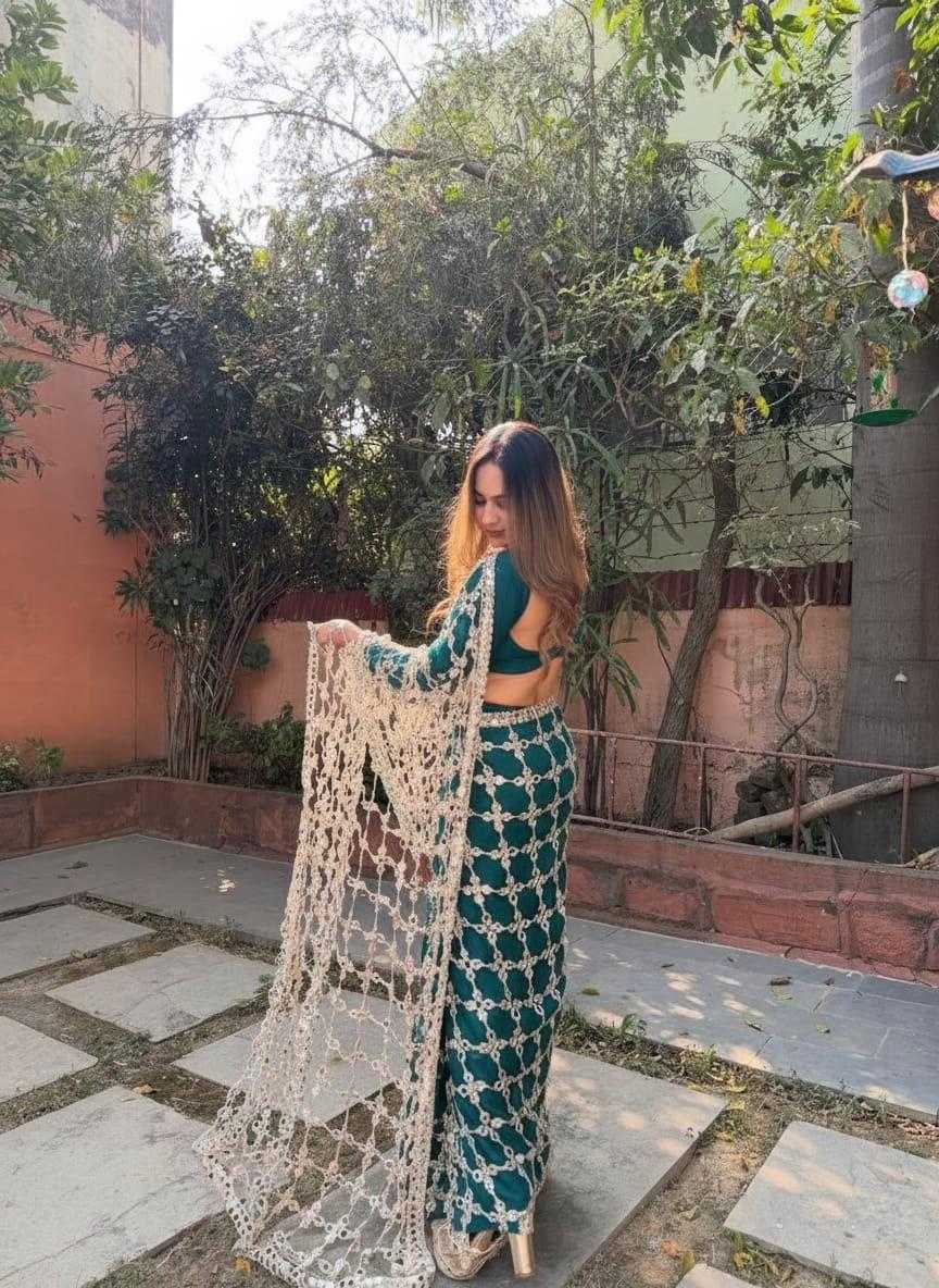 Woman in a green outfit with a white shawl standing outdoors near trees and a building.