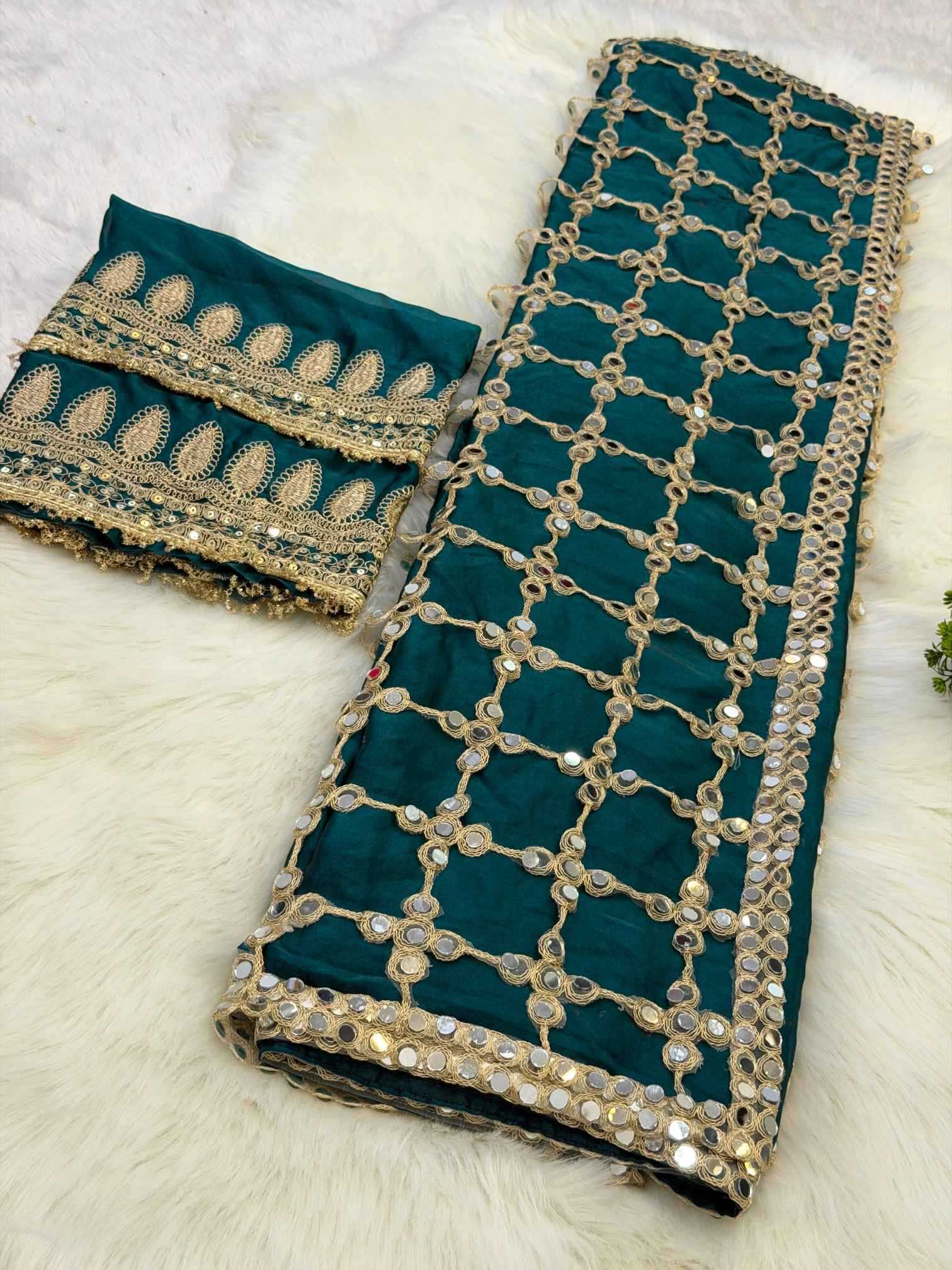 A folded dark teal green fabric with extensive gold zari embroidery, mirror work, and sequins, displayed alongside a matching embroidered border piece on a white faux fur background.