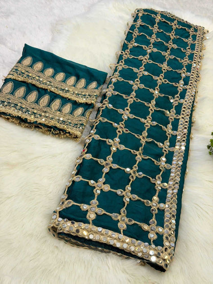 A folded dark teal green fabric with extensive gold zari embroidery, mirror work, and sequins, displayed alongside a matching embroidered border piece on a white faux fur background.