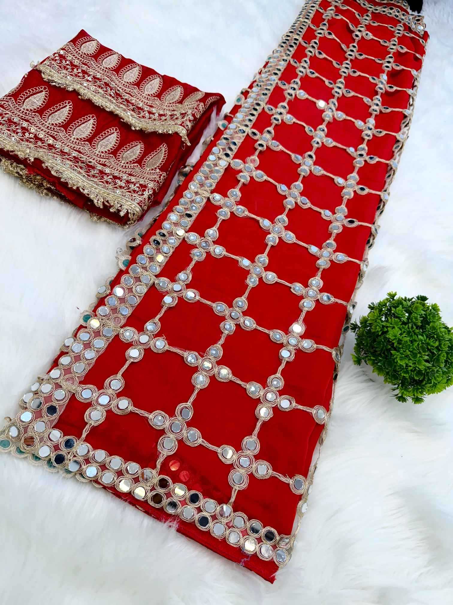 A red traditional embroidered fabric set with intricate silver mirror work and beige floral lace trim, displayed on a white faux fur background.
