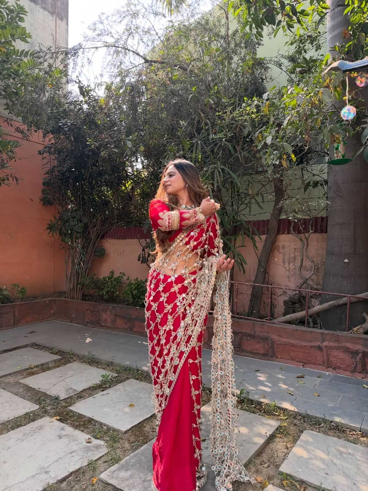 Woman in a red saree with gold details standing outdoors.