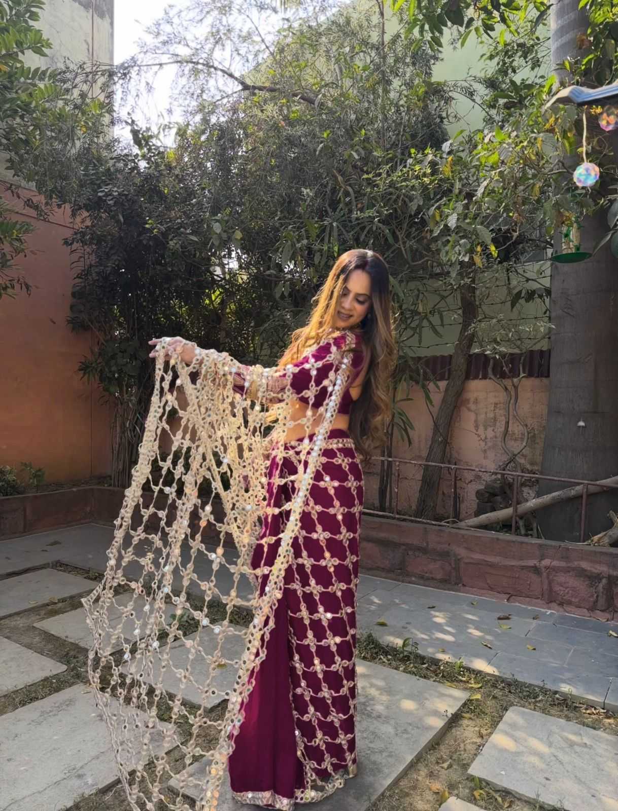 A woman stands outdoors in a garden courtyard wearing a deep magenta lehenga choli set with a matching sheer net dupatta. The outfit features intricate gold embroidery and a black waistband.