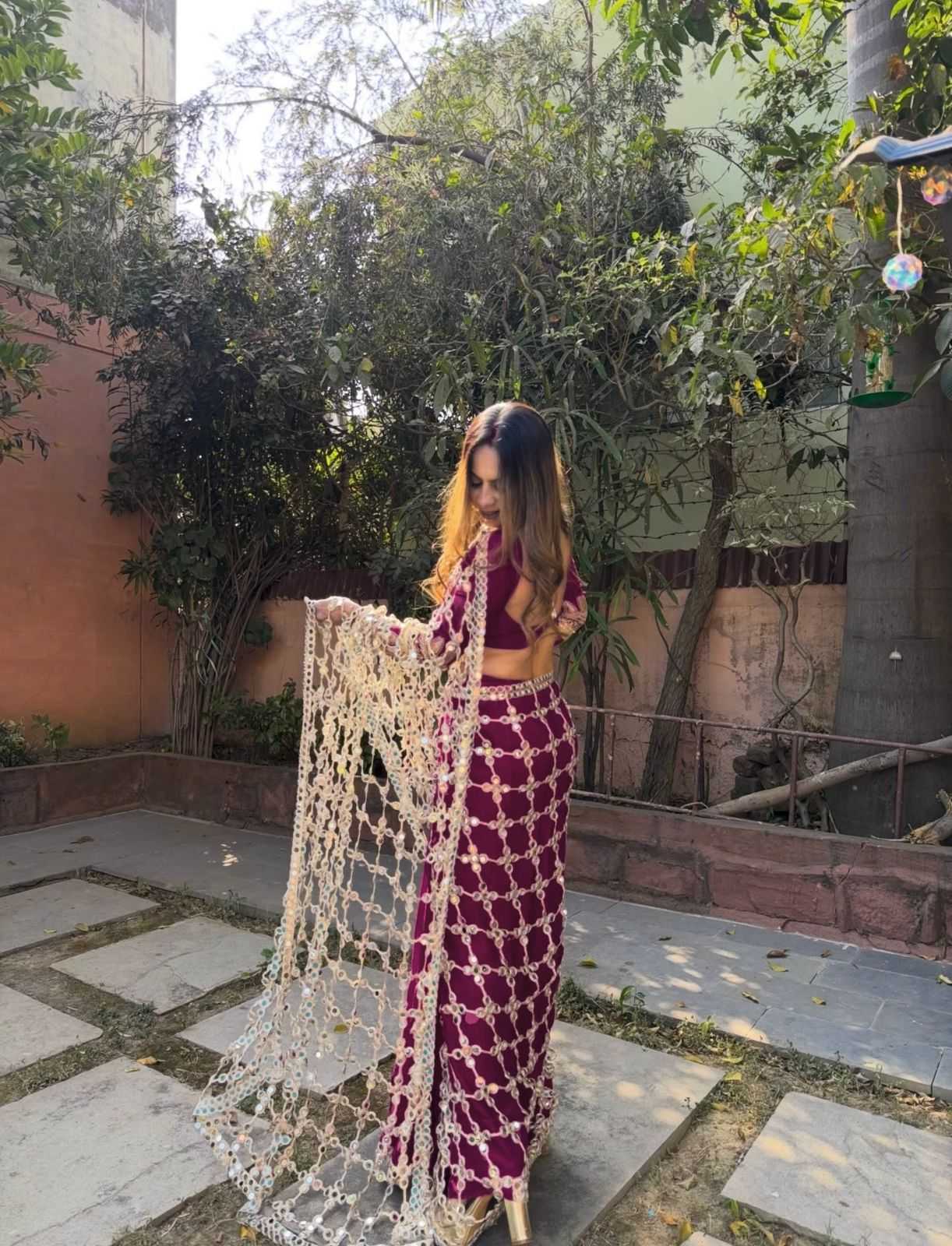 A woman stands outdoors in a courtyard wearing a deep purple embroidered traditional outfit with a matching long beaded scarf draped over one arm. The setting includes stone tiles, green trees, and a terracotta wall.