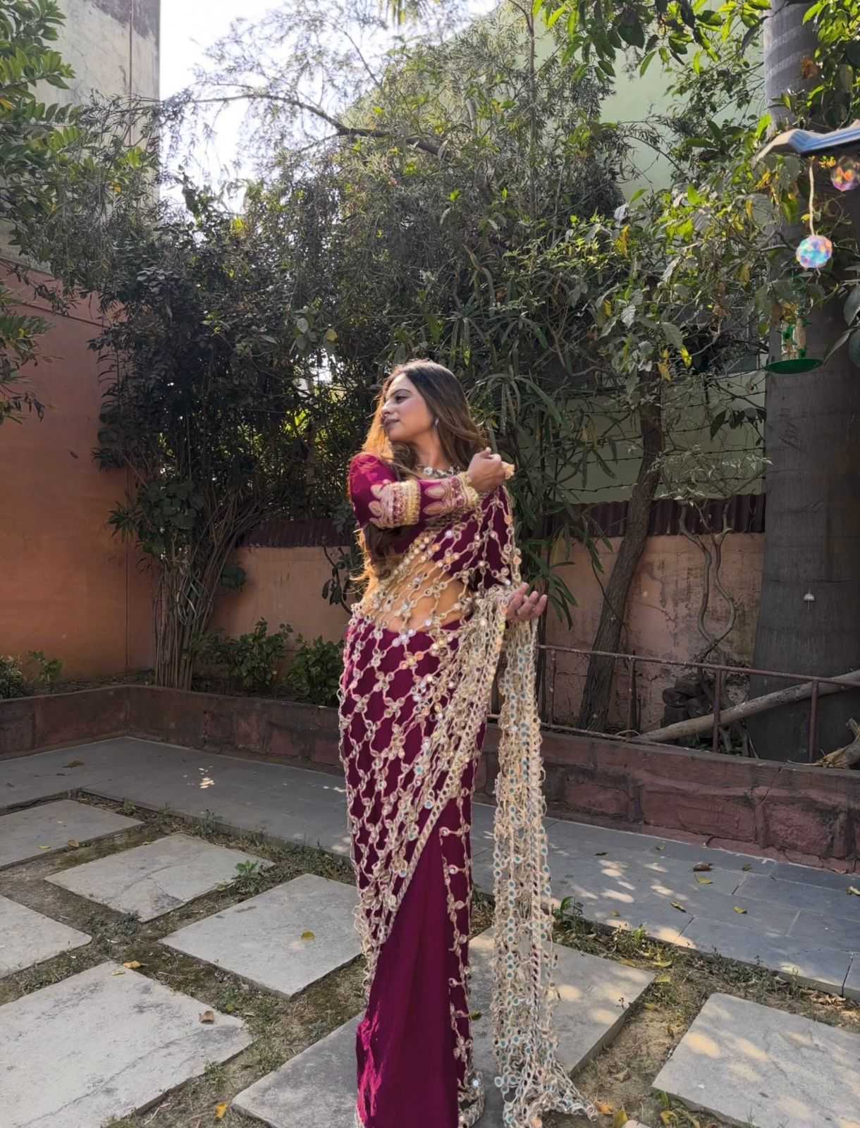 A woman wearing a traditional Indian outfit with a deep magenta skirt, a gold embroidered blouse, and a matching magenta and gold draped scarf, standing outdoors in a garden courtyard.