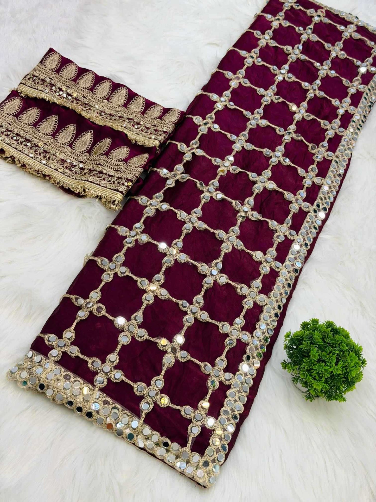 A deep maroon velvet saree fabric with intricate gold zari embroidery and mirror work, laid out on a white faux fur background next to a matching embroidered blouse piece and a small green plant.