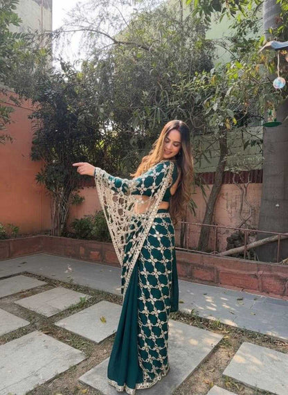 A woman wearing a dark teal green and gold embroidered traditional Indian outfit with a matching dupatta, standing outdoors on a stone patio with trees and a wall in the background.